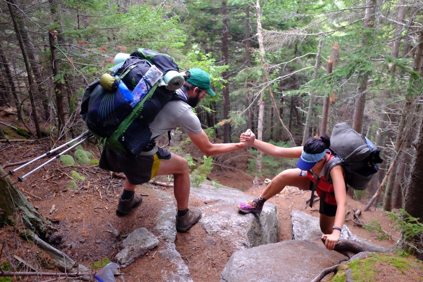 Two hikers on a forested trail, one giving a helping hand to the other as they navigate over large rocks. Both are carrying heavy backpacks and using trekking poles, indicating a challenging outdoor adventure.