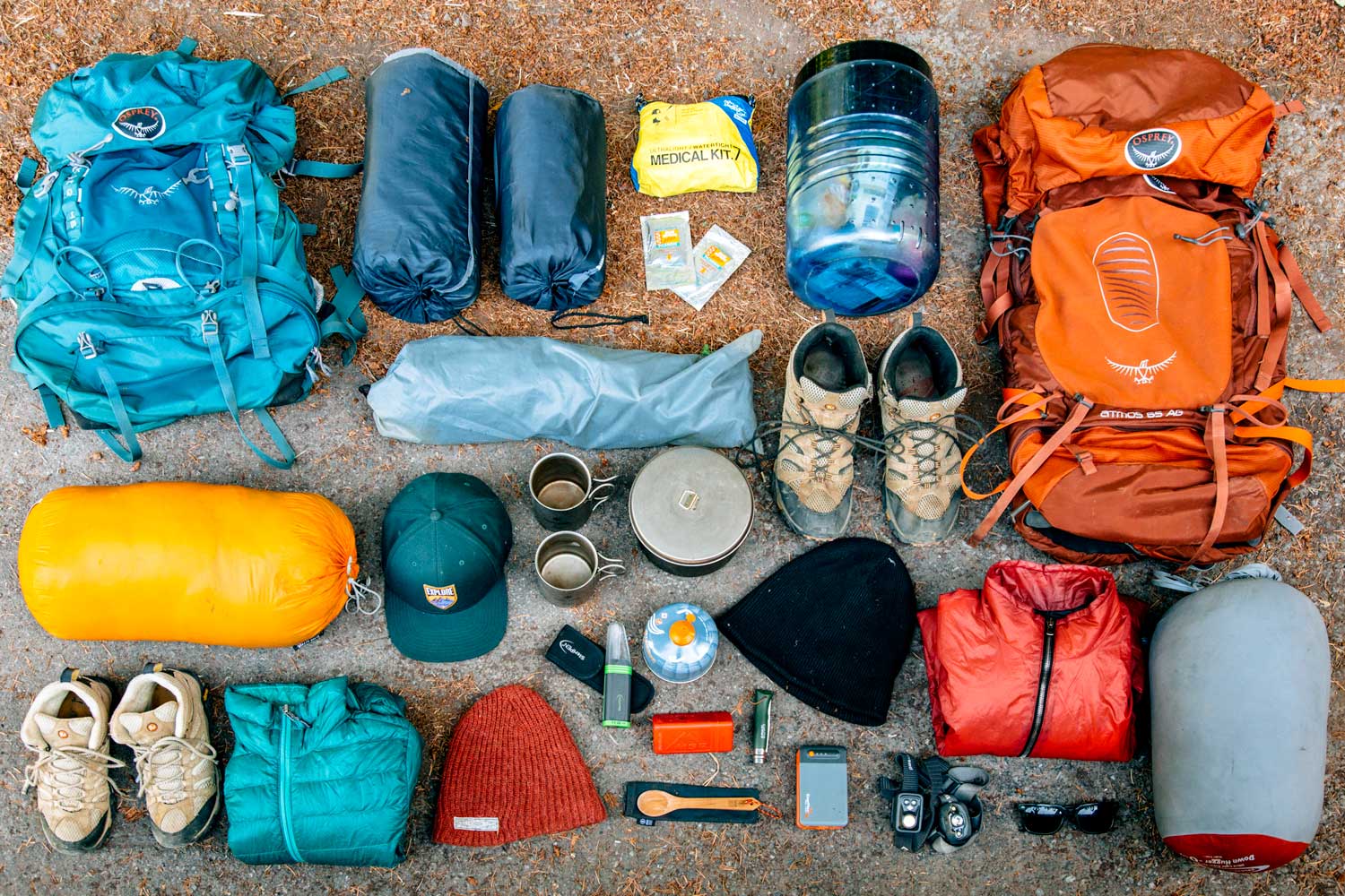 Outdoor gear neatly arranged on the ground, including two backpacks, a pair of hiking boots, a medical kit, a sleeping bag, cooking pots, a water bottle, a cap, a puffy jacket, a beanie, a flashlight, a portable GPS, a camera, and other hiking essentials.