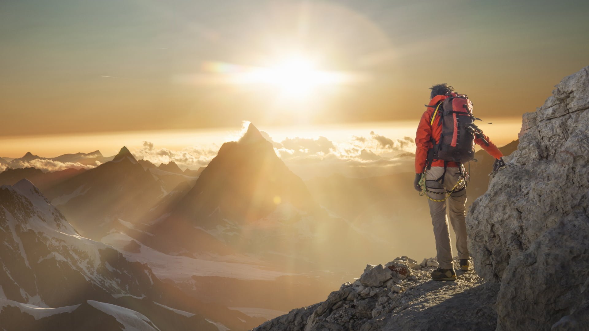 A hiker in orange gear stands on a mountain peak, overlooking a range of mountains bathed in the soft light of a sunset.